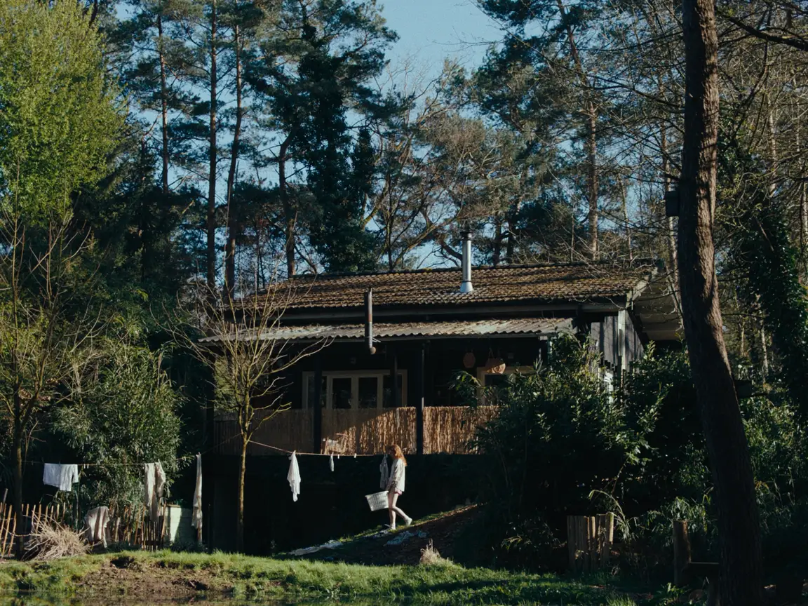 Moonlight Woman: Sarah carries a laundry basket past a wooden cabin among tall pines in sunlight