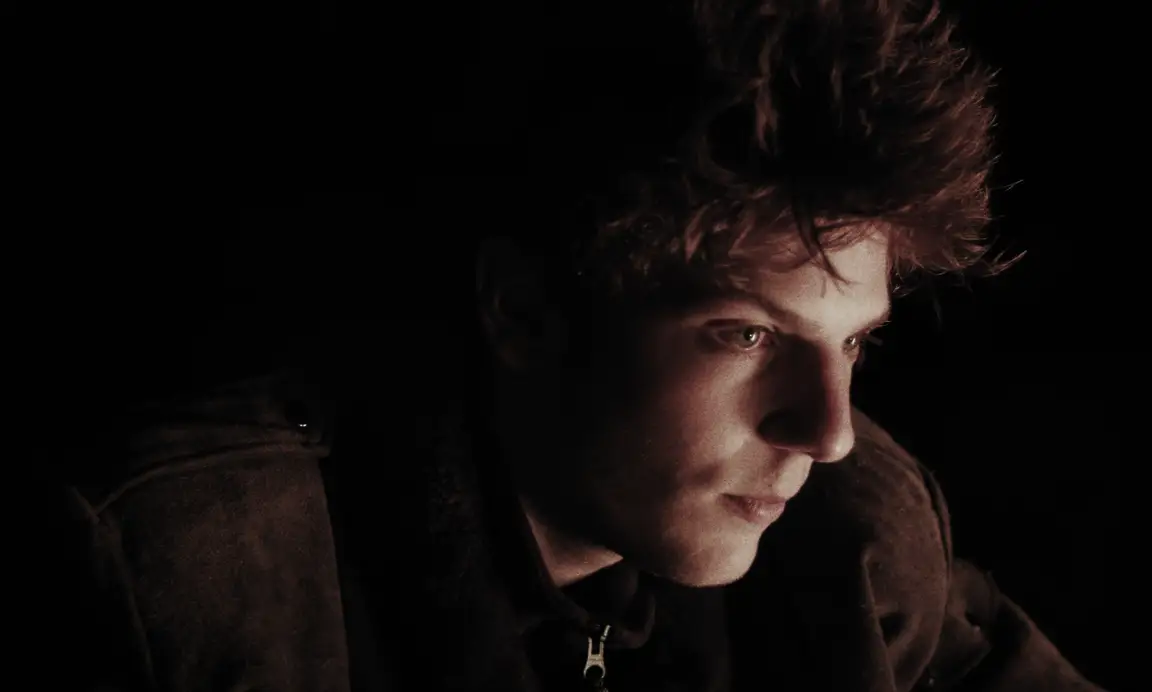 Felix, Le Gamin Qui Traverse: curly-haired young man in warm low light looks off into the surrounding darkness