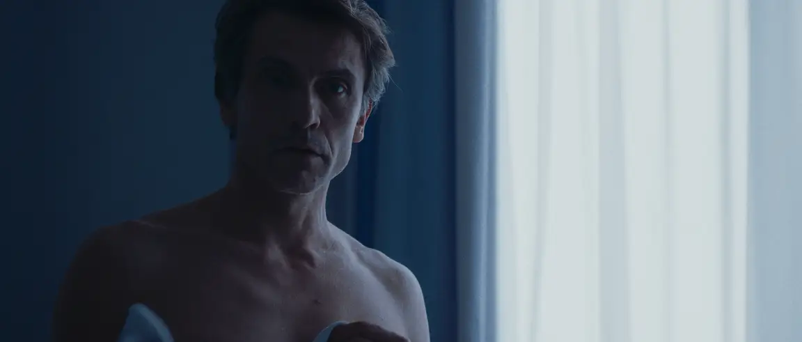 La Belle Rosine: shirtless professor lit by cold blue light beside a sheer curtained window