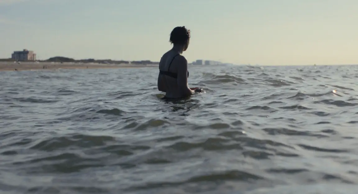 Burn: a woman stands waist deep in the sea facing the horizon, coastline behind her