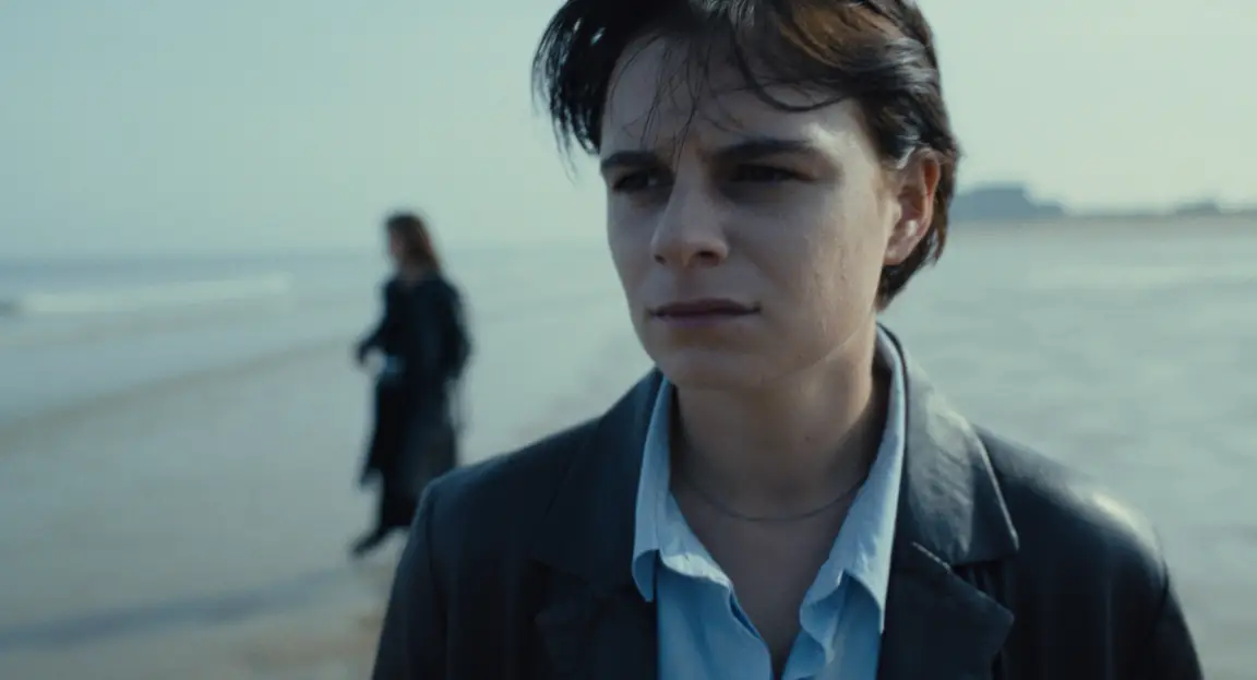 Burn: a short-haired woman in a leather jacket on the beach, a dark figure behind her