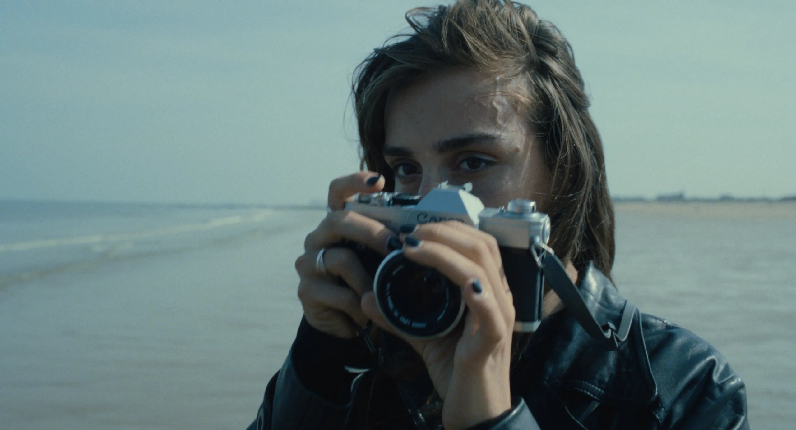 Burn: a woman raises a vintage Canon camera to her eye on an overcast beach