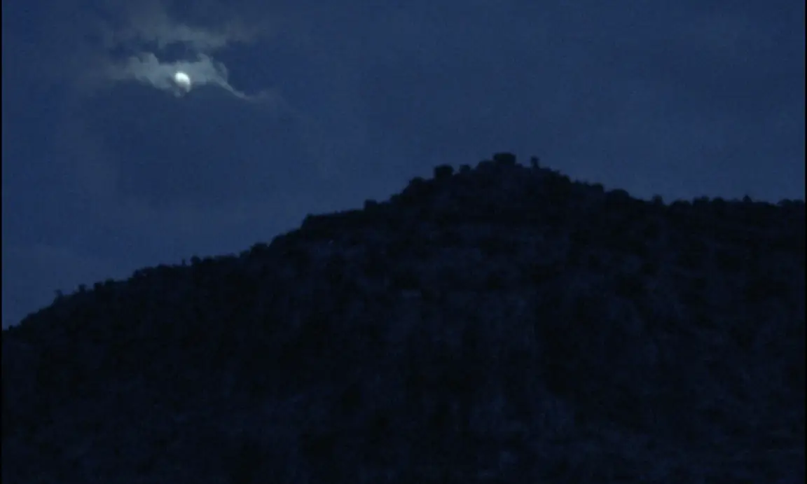 Ever Since, I Have Been Flying: the moon breaks through cloud above a dark silhouetted hilltop at night