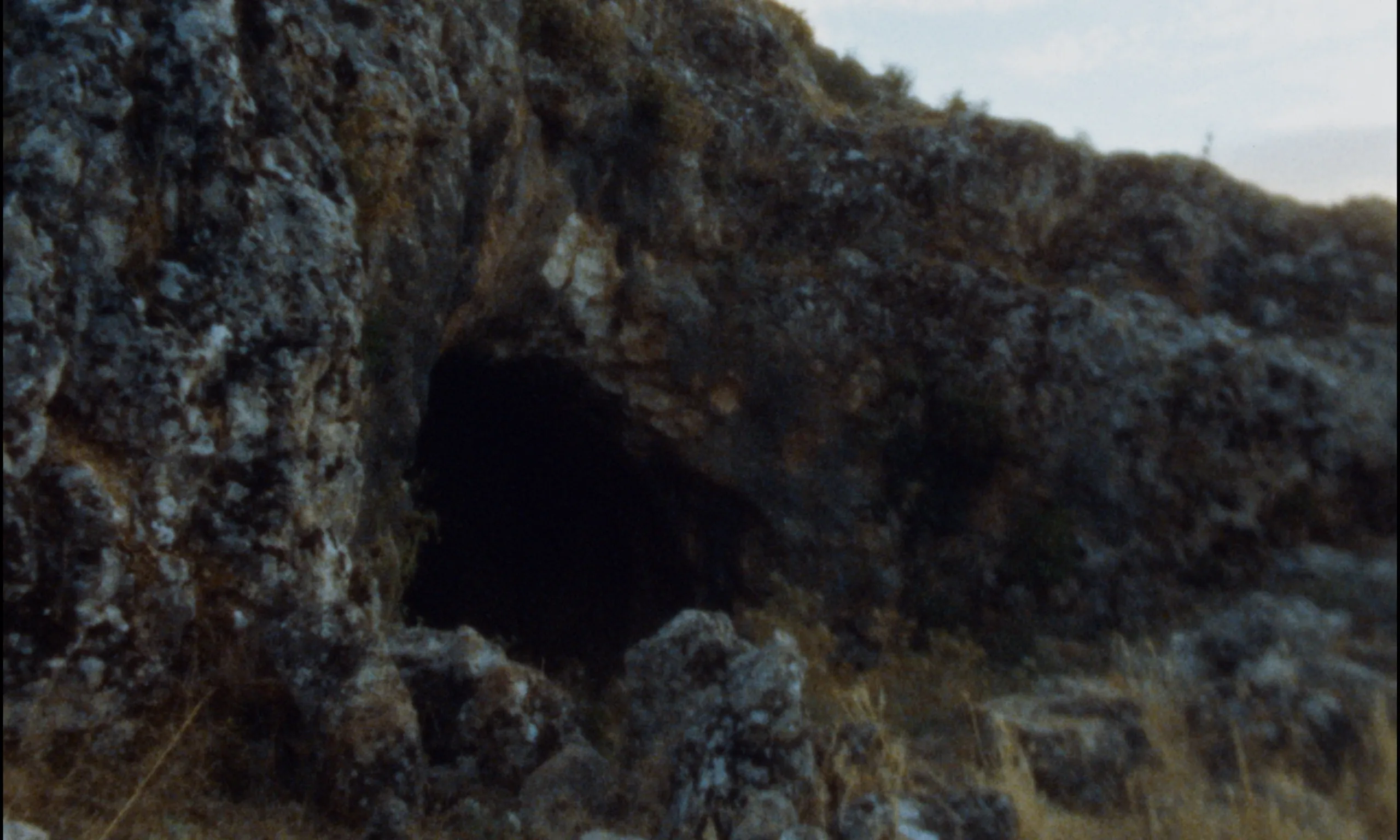 Ever Since, I Have Been Flying: a dark cave mouth set into a rugged rocky outcrop under a pale sky