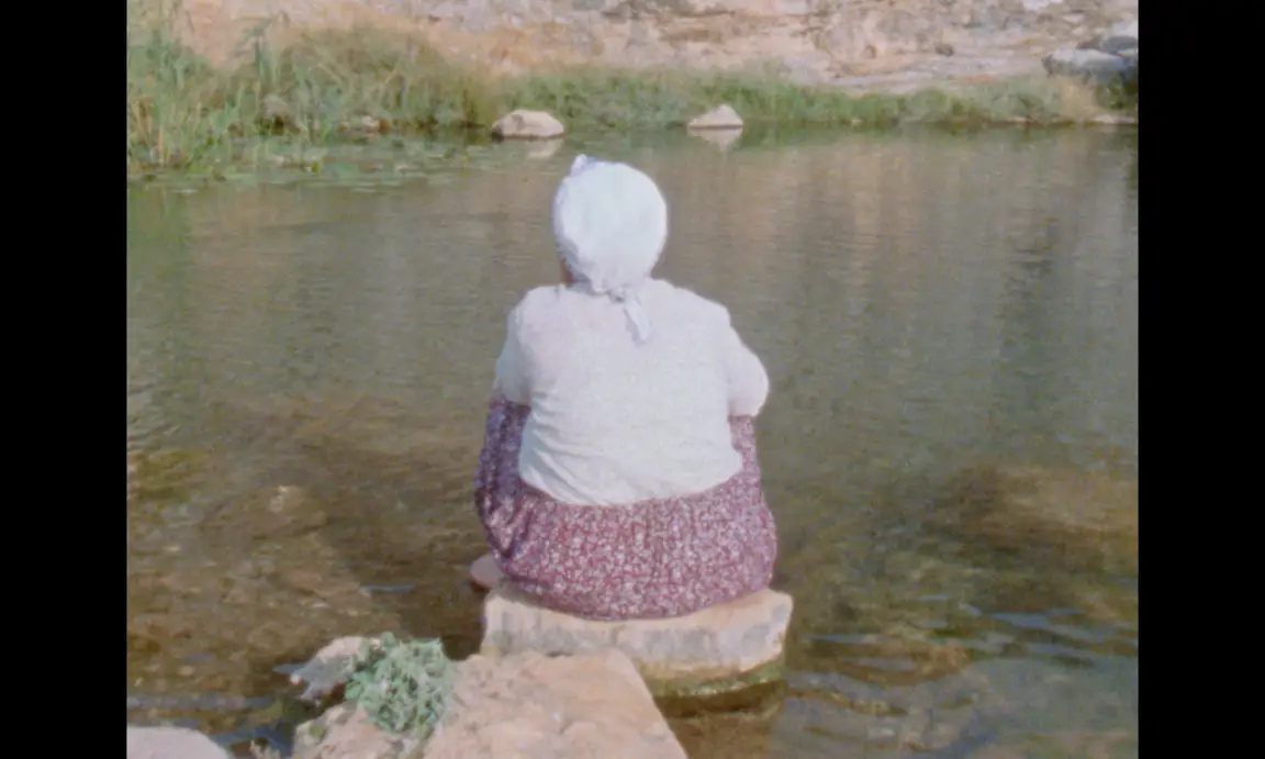 Ever Since, I Have Been Flying: an older woman in a headscarf sits from behind on a rock at the water edge