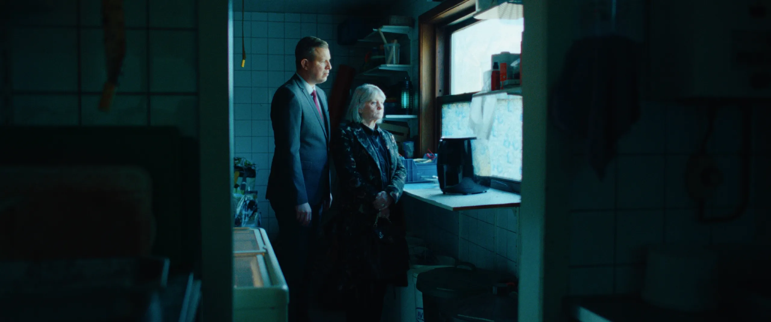 Vlinderman: a suited man and an older woman wait in a cramped tiled kitchen