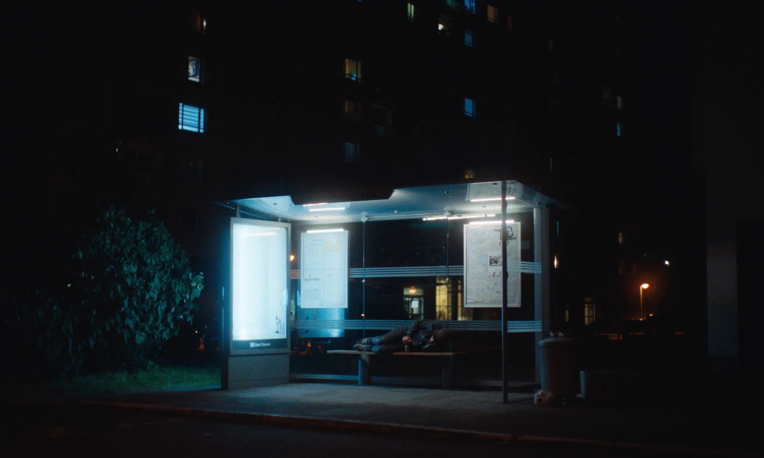 Burning Clouds: a man lies asleep on the bench of a glowing bus shelter at night