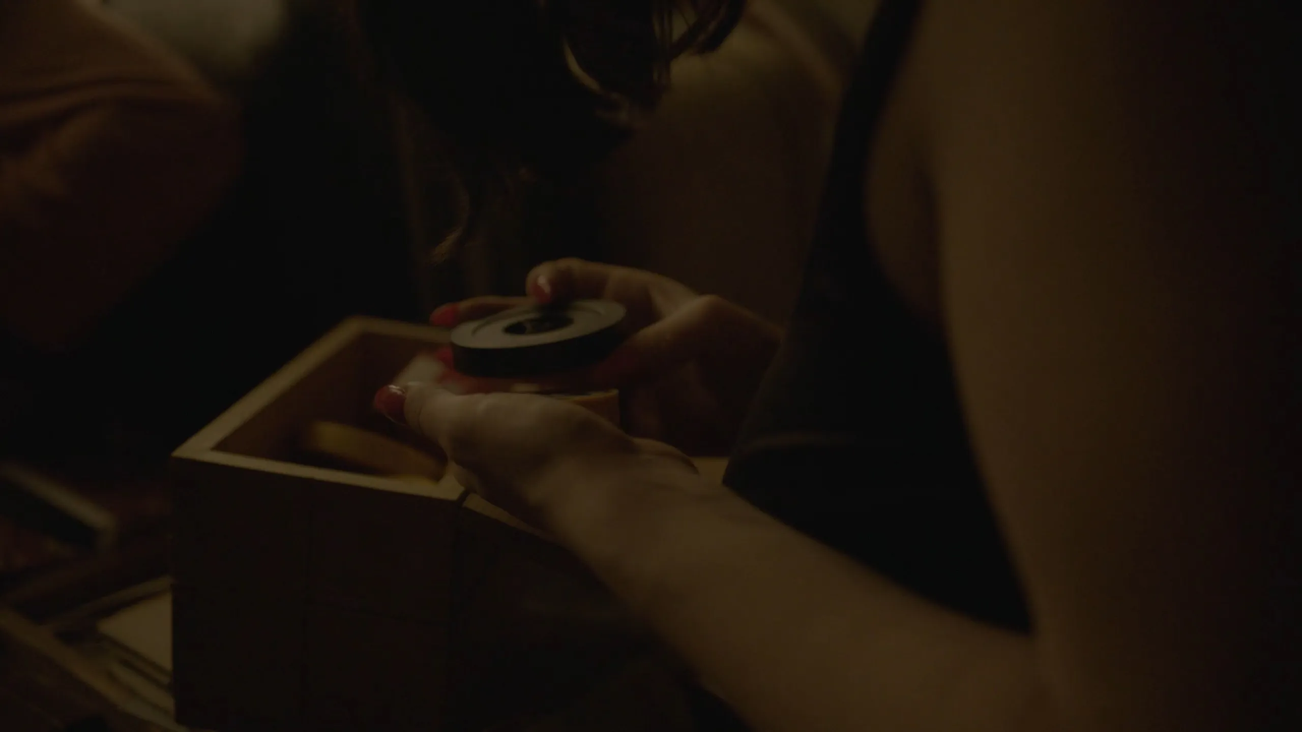 Chance Encounters: red-nailed hands lifting a film canister from a wooden box in low warm light