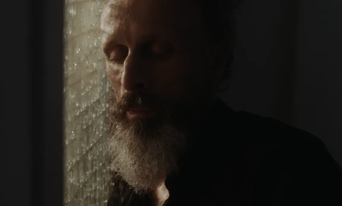 Les Homards Immortels: bearded man closes his eyes beside a textured glass window in soft light
