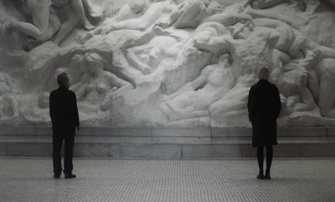 Les Homards Immortels: a man and a woman stand apart before a monumental marble relief of entwined bodies