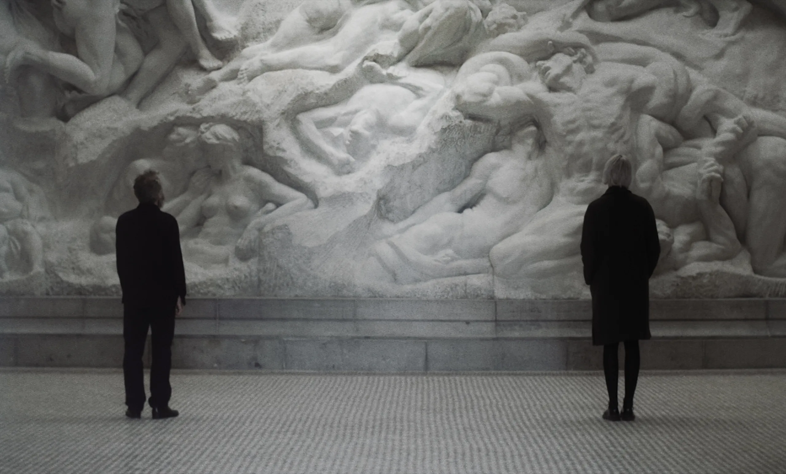 Les Homards Immortels: a man and a woman stand apart before a monumental marble relief of entwined bodies