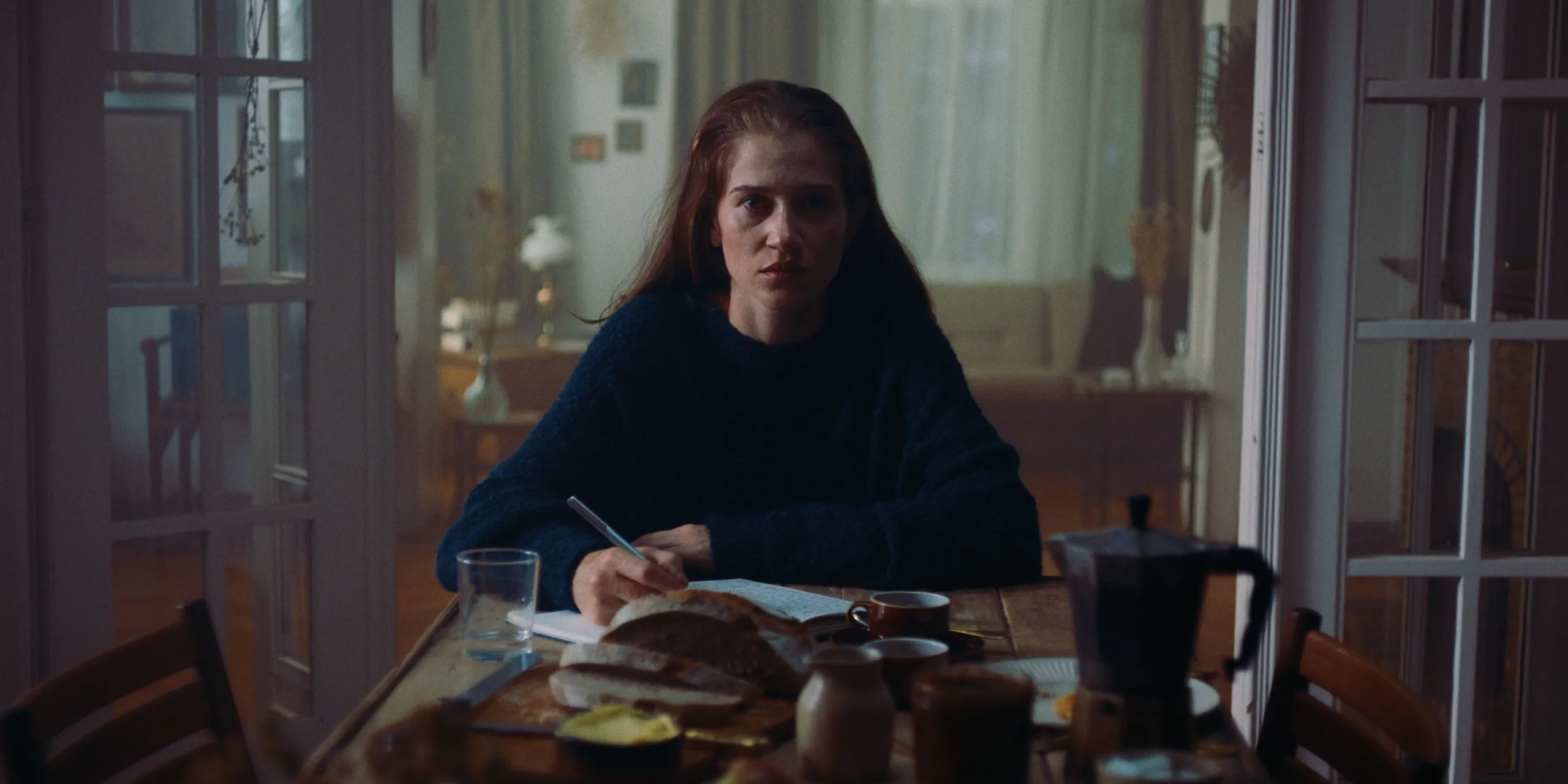 The Tears of Things: a woman in a blue sweater writes in a notebook at a breakfast table