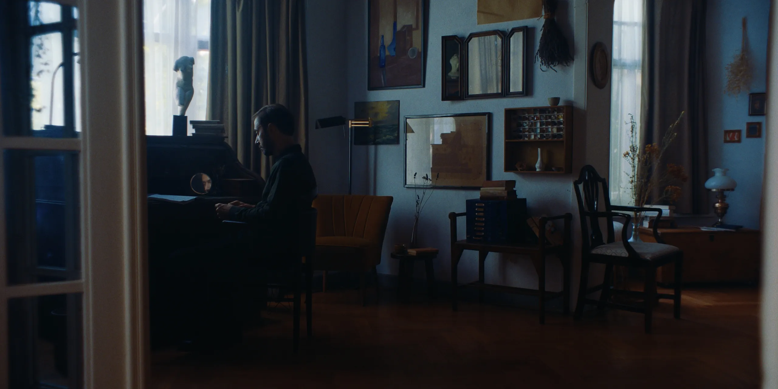 The Tears of Things: a man sits playing an upright piano in a cluttered art-filled living room