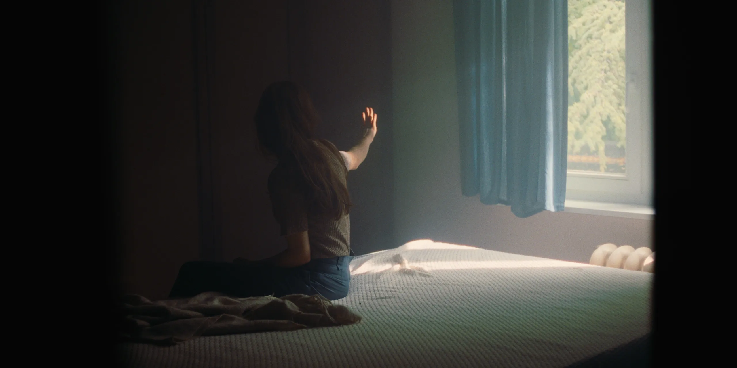 The Tears of Things: a woman kneels on a bare mattress reaching toward a sunlit curtained window