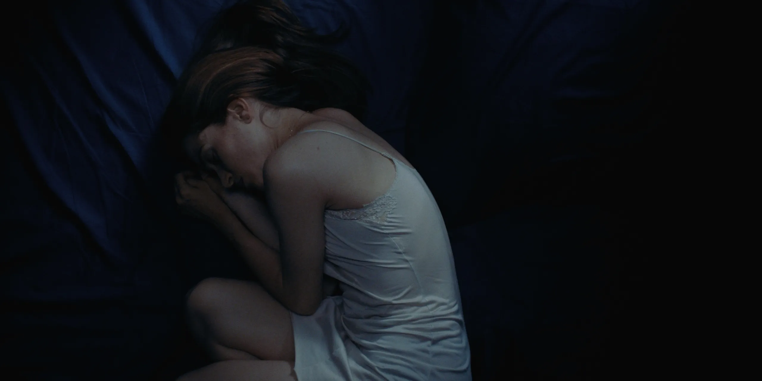 The Tears of Things: a woman in a white slip curls up on dark bedding in shadow