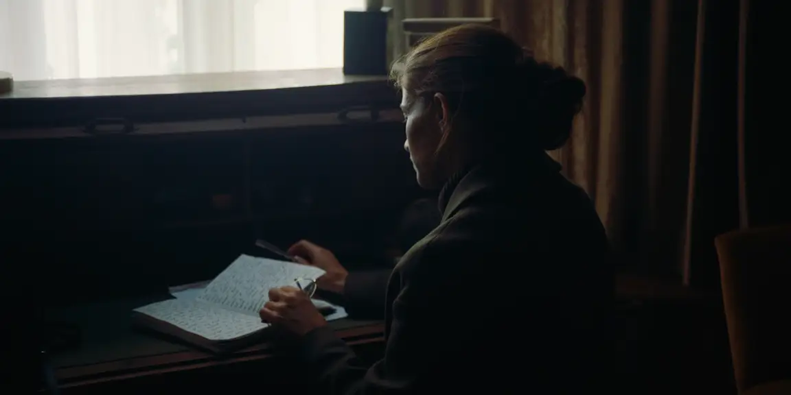The Tears of Things: a woman sits at a desk writing in her diary by a bright curtained window
