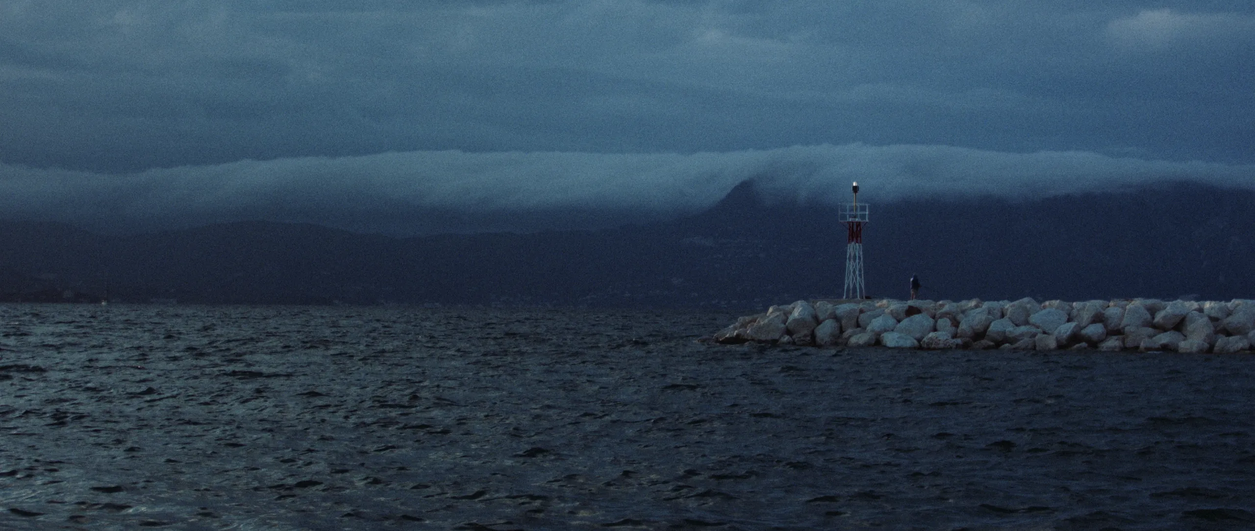 Springtide: a lone figure stands by a small beacon tower on a rocky breakwater at dusk