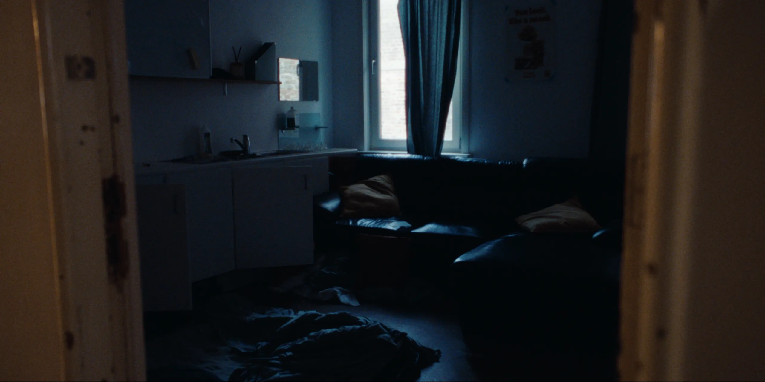 Bearer of Bad News: dim blue apartment room with kitchenette, leather sofa and bedding strewn on the floor
