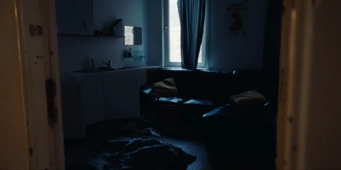 Bearer of Bad News: dim blue apartment room with kitchenette, leather sofa and bedding strewn on the floor