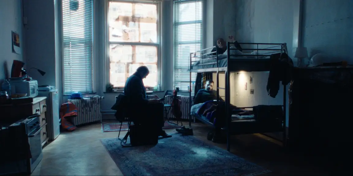 Bearer of Bad News: Sven sits in a migrant workers dorm with bunk beds and newspapered windows