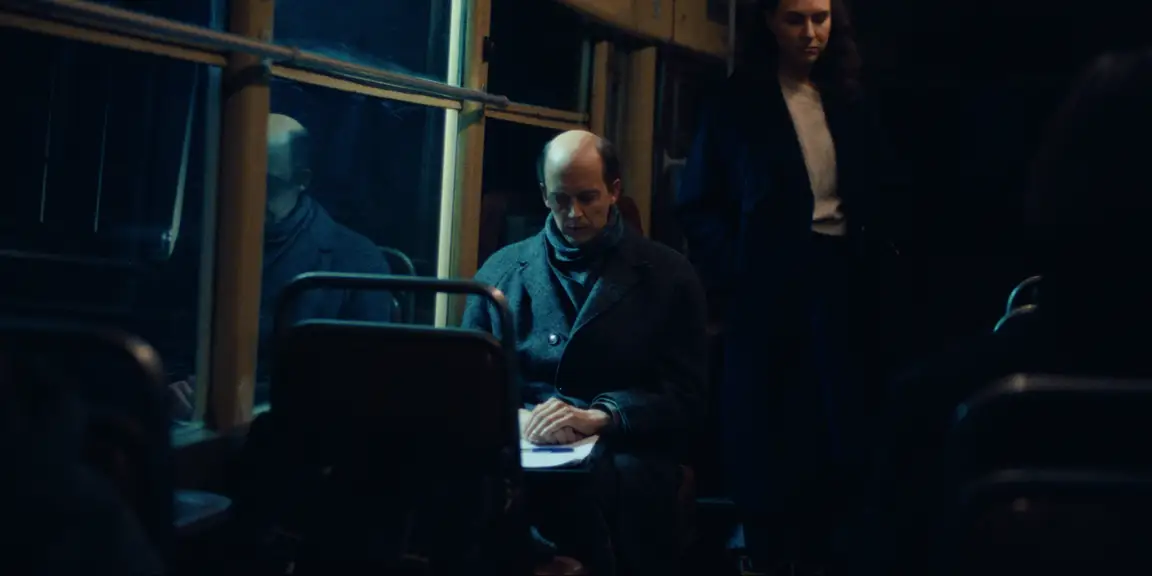 Bearer of Bad News: seated on the tram, Sven folds his hands over papers while a woman watches