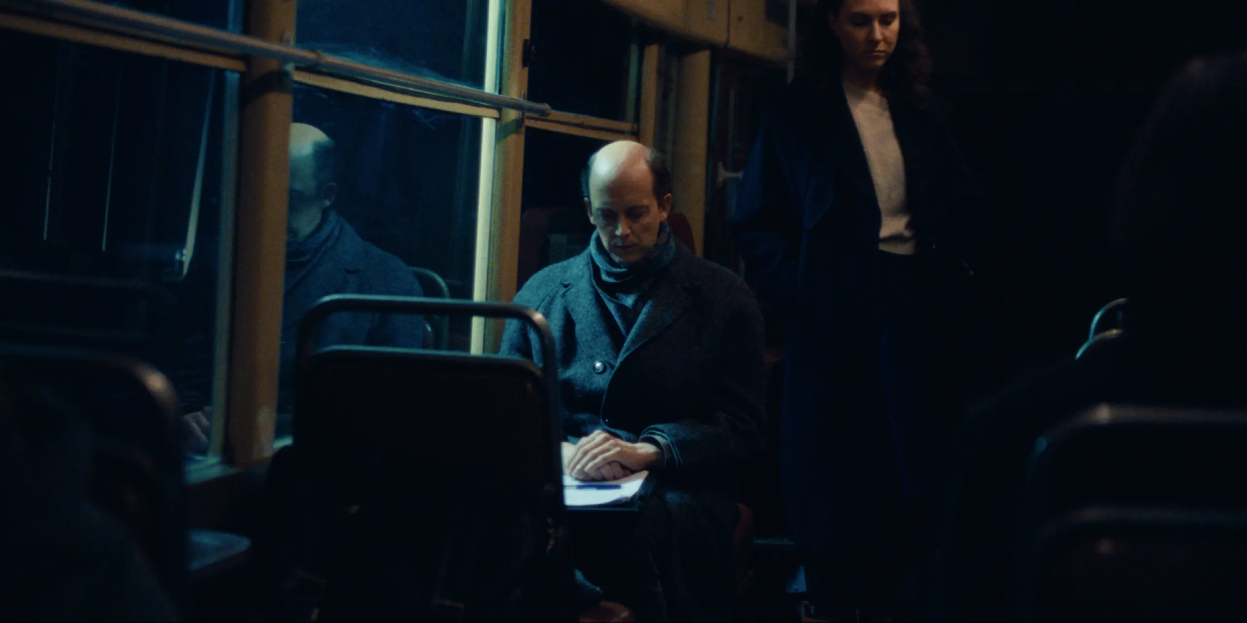 Bearer of Bad News: seated on the tram, Sven folds his hands over papers while a woman watches