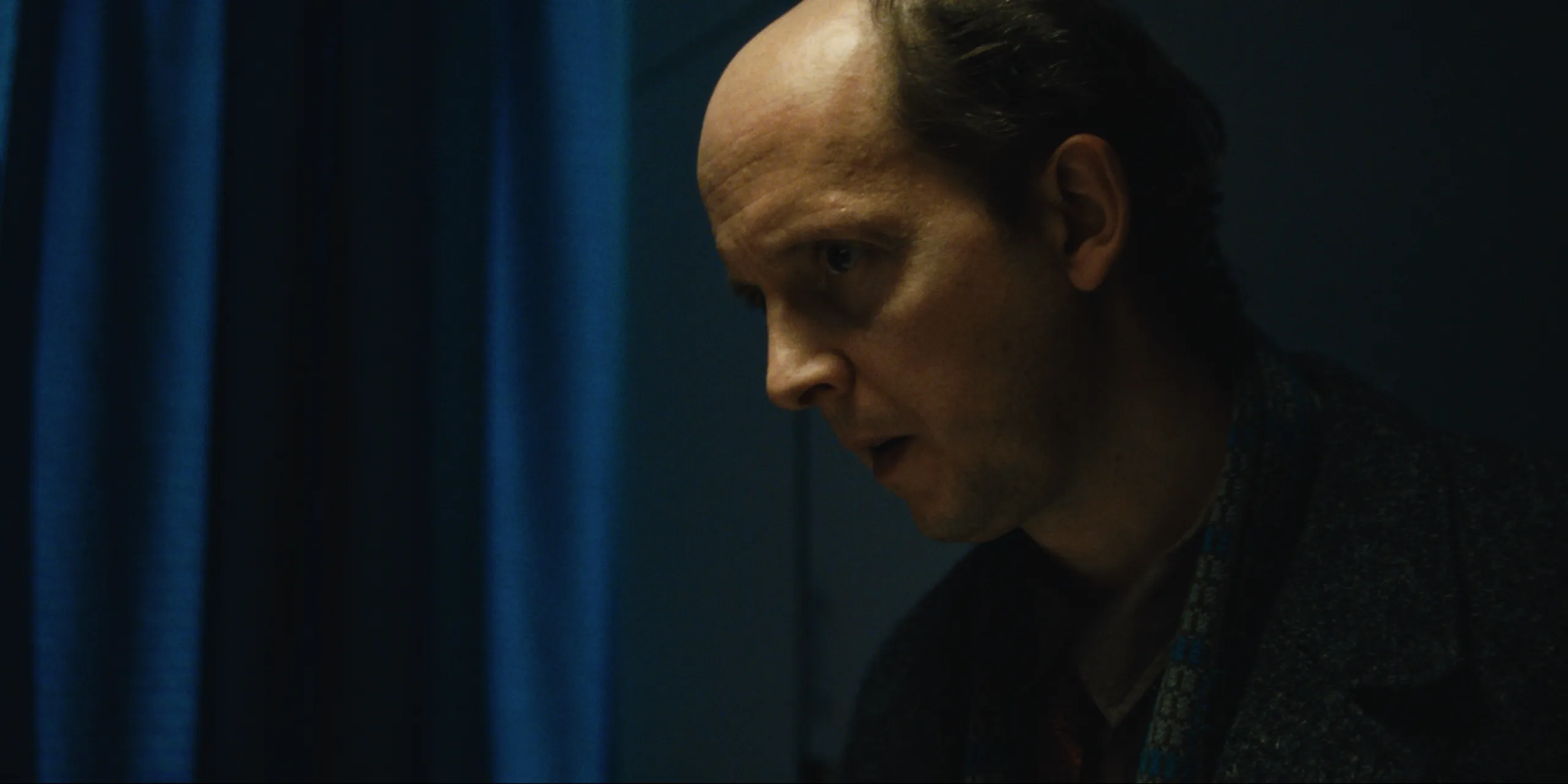 Bearer of Bad News: profile of Sven looking down beside a blue hospital curtain in dim light