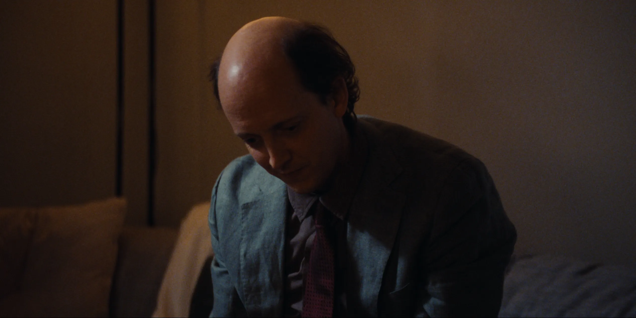 Bearer of Bad News: Sven sits on a sofa edge in suit and tie, head bowed in lamplight