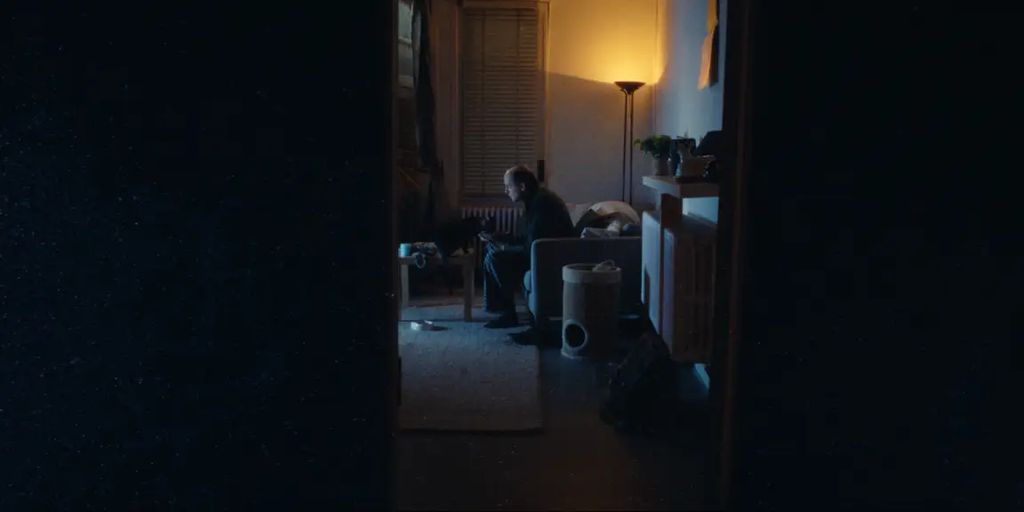 Bearer of Bad News: seen through a dark doorway, Sven sits with his cat by a warm floor lamp