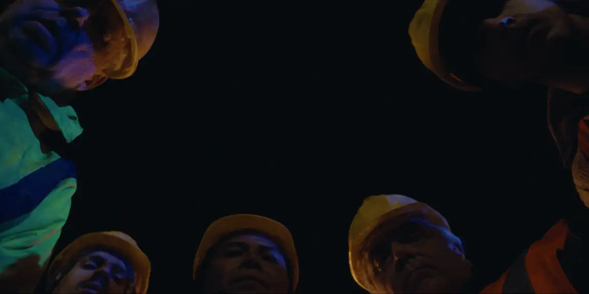 Bearer of Bad News: ring of construction workers in yellow hard hats looking down from above at night