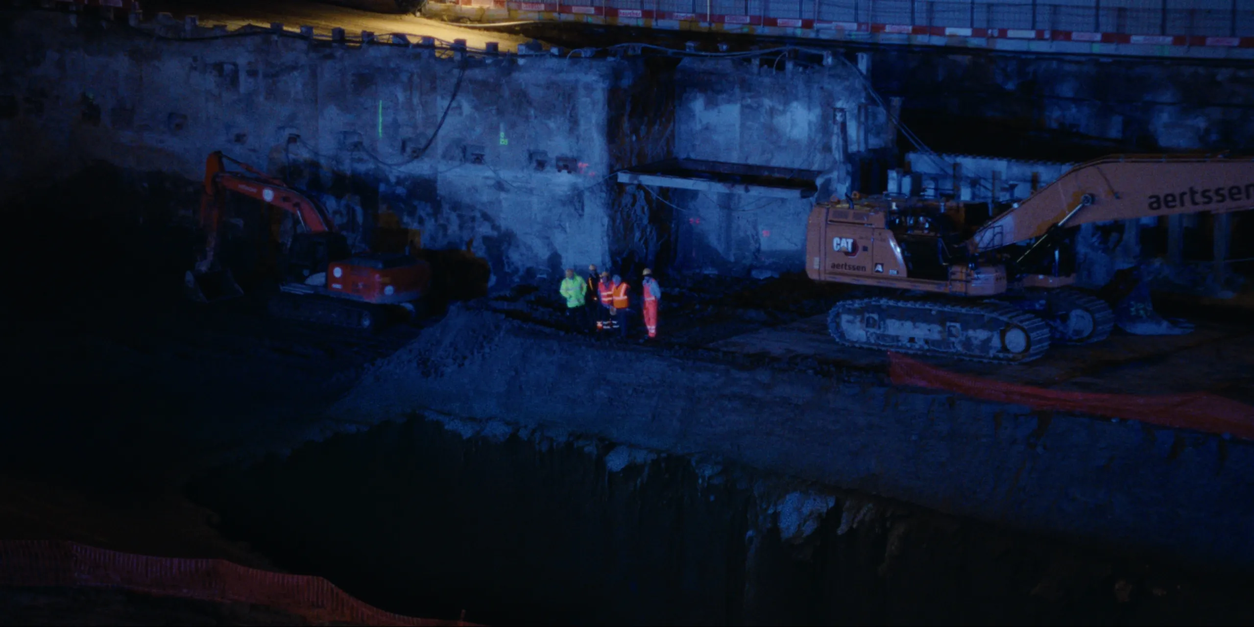 Bearer of Bad News: floodlit construction pit at night with excavators and workers in hi-vis on a ledge