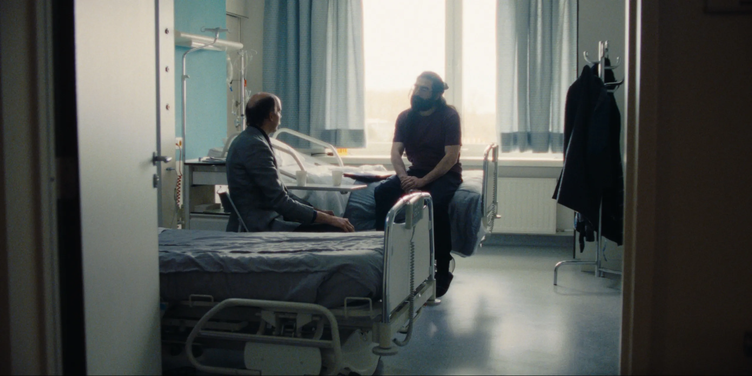 Bearer of Bad News: Sven sits beside a hospital bed talking with a bearded patient by a bright window